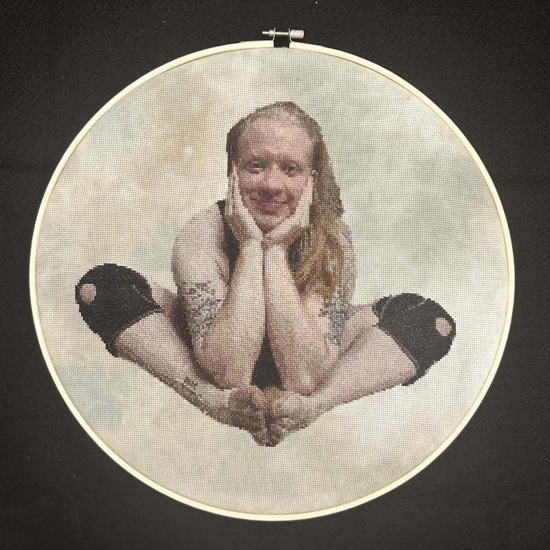 A cross-stitch portrait in a large circular embroidery hoop of a white person with long strawberry blonde hair, sitting bent over with their feet touching and elbows on their feet, with their hands cupping their face. They are looking at the camera with a slight smile, and have visible tattoos on both biceps. They are wearing black knee braces on both knees and an ankle brace on their right ankle.