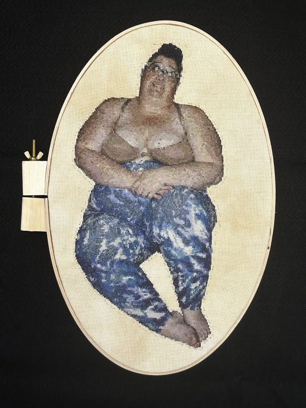Image Description: A cross-stitch portrait of a fat person sitting with their hands crossed over their belly and knees bent, looking off to the left. They are wearing a tan bra and tie dye leggings, have light skin with red patches, and have dark hair in a high bun. 