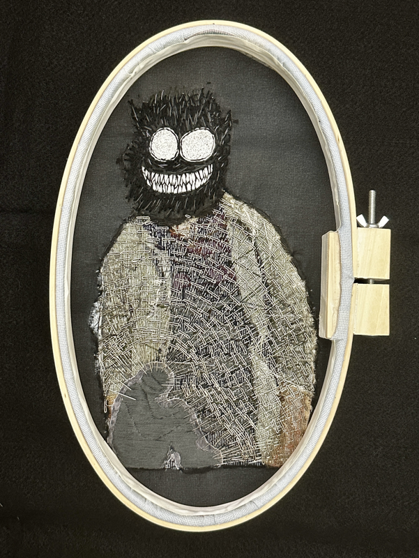 The reverse side of the piece shows the figure with an embroidered black grinning monster head with large pointed white teeth and enormous circular white eyes. The body of the figure has a silhouette of a small huddled person with a sideways cap embroidered in the bottom left corner, and the rest of the body is filled with T-pins, encircling and piercing the figure. The background is covered in a sheer black fabric and is tearing around the edges. 