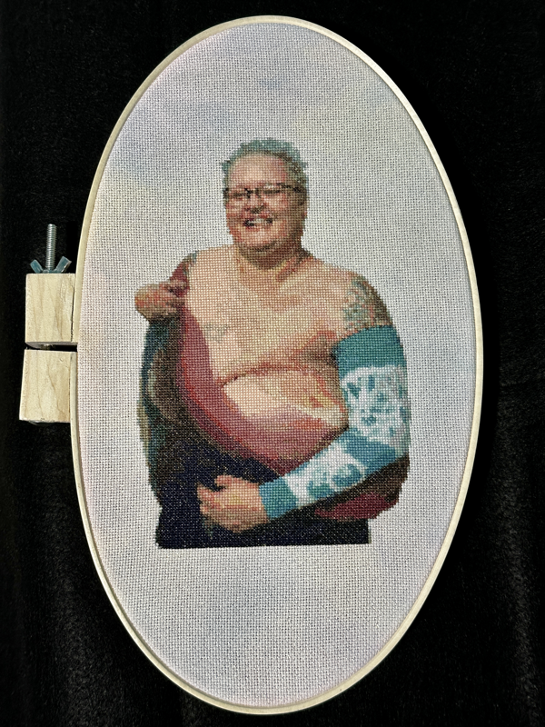 A cross-stitched portrait of a laughing white woman with very short teal hair and glasses. She is topless and holding a rose-coloured cloth to cover one breast, the exposed side of her chest has red mastectomy scars where the other breast has been removed. She is wearing a teal arm sleeve, from which a part of a large, colourful tattoo can be seen poking out the top, and a dark skirt. 