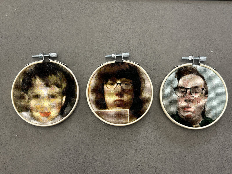 Three small circular cross-stitch portraits in embroidery hoops. The first portrait (left) is of a toddler with light brown hair and very orange lips. The middle portrait shows a young person with medium length brown hair and glasses, holding a piece of paper to the camera with a somber face. The final portrait (right) features a non-binary person with short spiky brown hair, wearing glasses and a green collared shirt. 