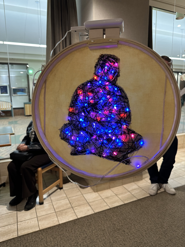 The reverse side of the piece shows the figure covered in knotted and bunched small LED lights, wrapped tightly with black criss-crossing lines. The lights are in a variety of colours, but mostly red, blue, and purple.