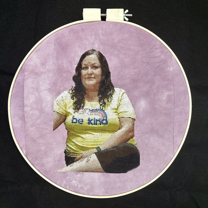 A cross-stitch portrait of an Indigenous woman with long dark hair, sitting with one leg crossed and a hand on her tattooed calf. She is looking at the viewer and wearing a yellow t-shirt that says “Be Kind” with a rainbow. The background is mottled mauve colour. 